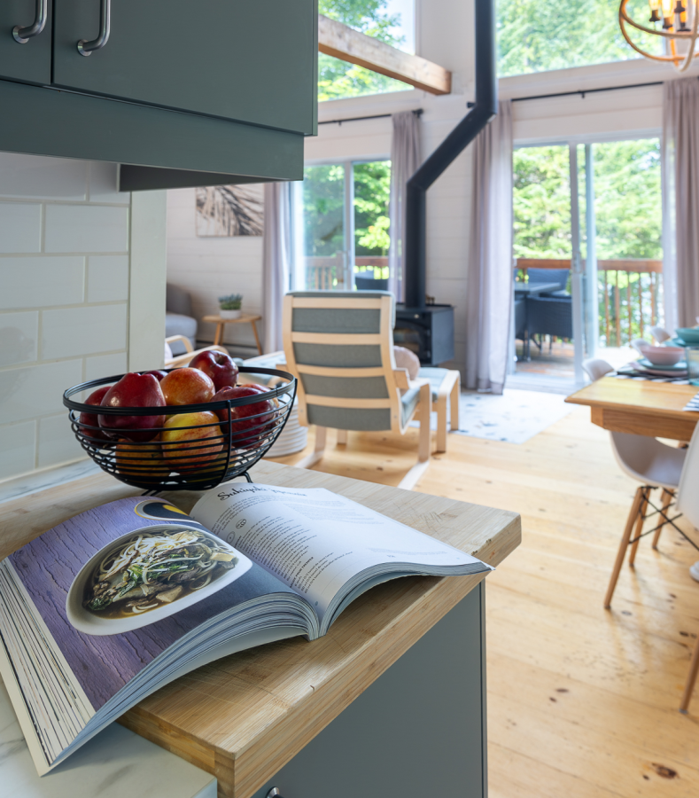 Le Mandala’s kitchen open to the living room, a cookbook and a bowl of apples on the island, wood stove and patio doors beyond