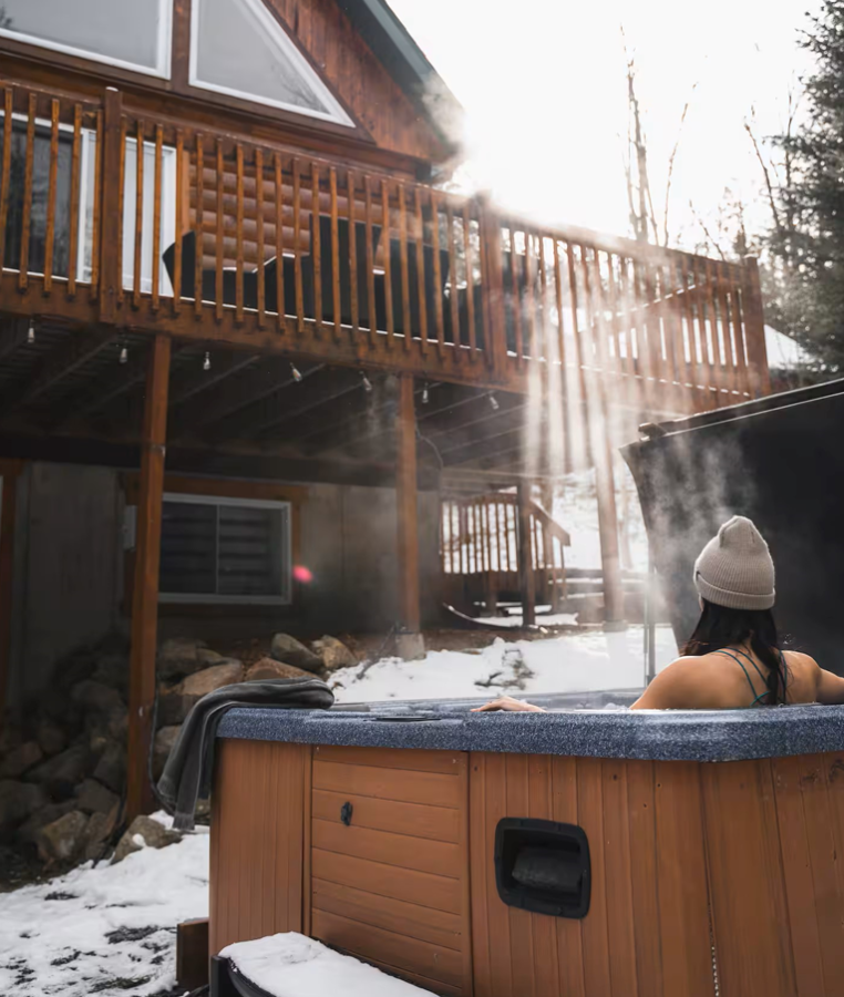 Steaming outdoor hot tub in the snow, someone in a toque in the water, a large wood deck and low sun behind the trees