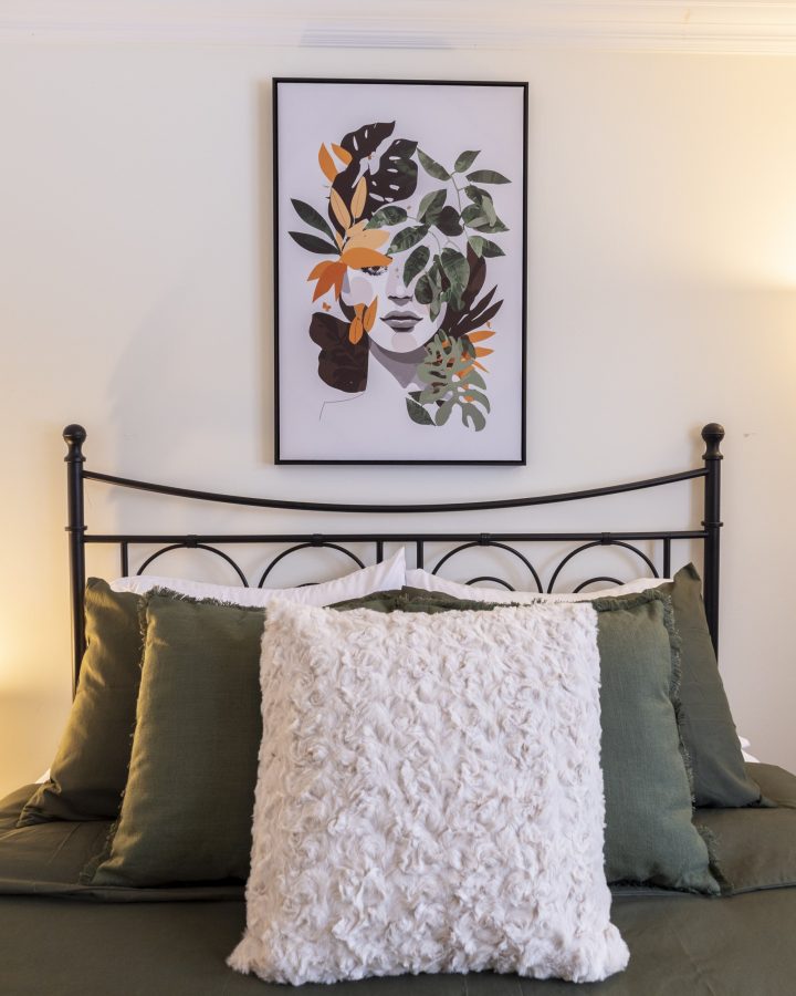 Un lit en fer forgé noir, coussins de lin vert sauge et coussin de fourrure crème, sous une affiche encadrée de feuillages.